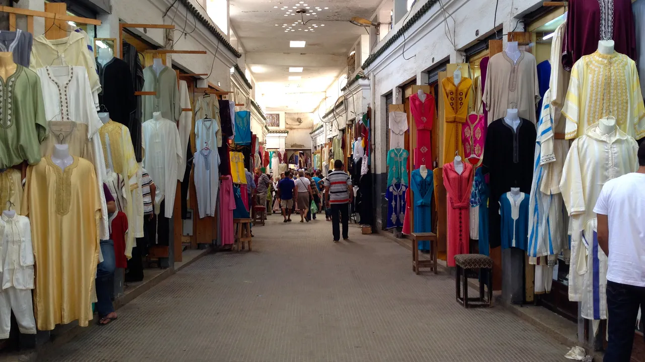 Allée commerçante de la médina de Fès bordée de boutiques de djellabas et de caftans, avec des passants