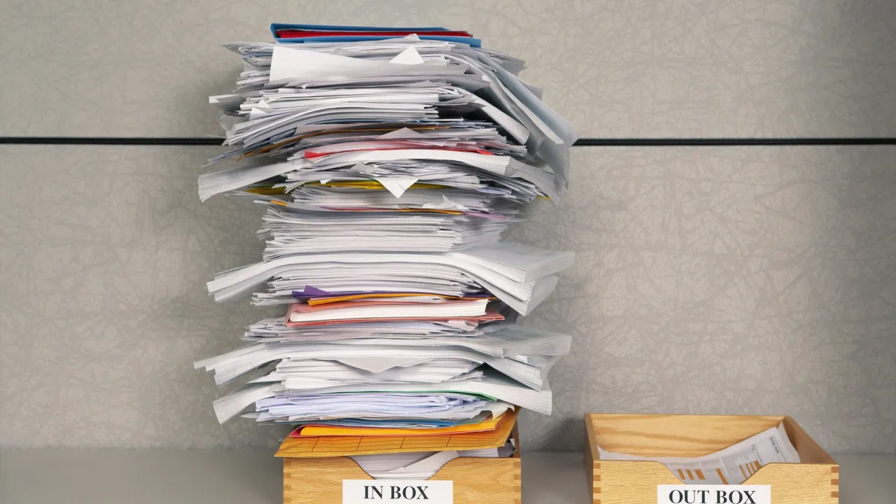 Pile de documents administratifs dans une corbeille « In box », à côté d'une corbeille « Out box » vide